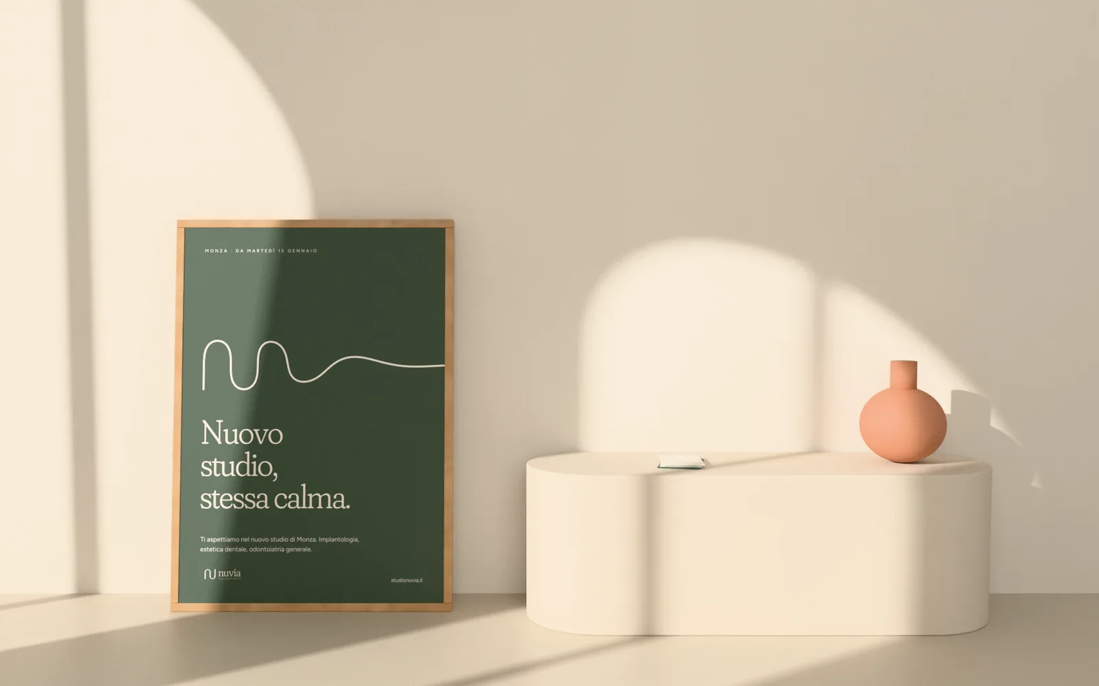 Manifesto verde salvia della campagna di apertura Nuvia con il filo color avorio e il titolo Nuovo studio, stessa calma, in cornice di rovere appoggiato a una parete illuminata da finestre ad arco